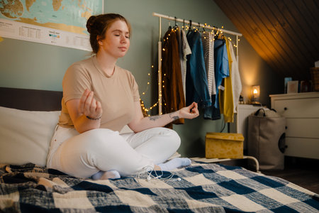 Young beautiful girl meditating with closed eyes in lotus pose on bed in cozy room at homeの写真素材