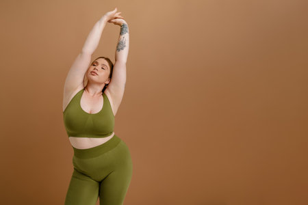 Young beautiful calm plus size girl in training suit stretching her arms with closed eyes , while standing over isolated brown backgroundの写真素材