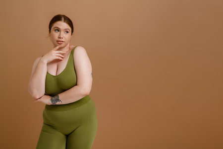 Young beautiful calm plus size girl in training suit touching her chin and looking aside , while standing over isolated brown backgroundの写真素材