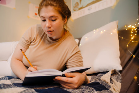 Young beautiful focused girl writing in notebook with pencil , while lying on bed in cozy room at homeの写真素材