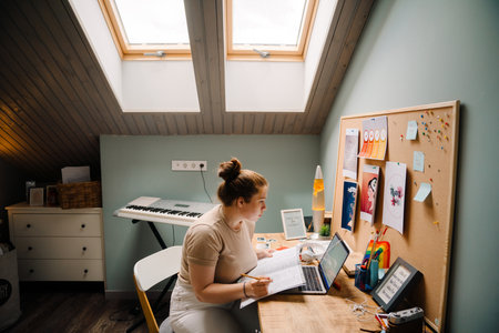 Young beautiful focused girl holding pencil looking on laptop, while sitting in cozy room at homeの写真素材