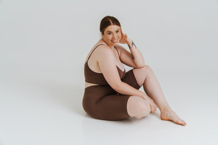 European woman with ginger hair smiling while sitting on floor isolated over white backgroundの写真素材