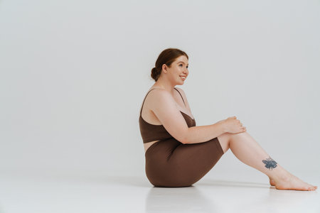 European woman with ginger hair smiling while sitting on floor isolated over white backgroundの写真素材