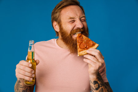 Bearded happy man holding pizza slice and beer while standing isolated over blue backgroundの写真素材