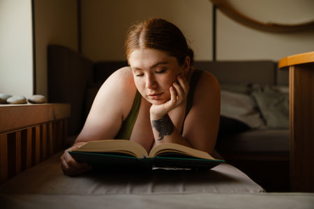 Ginger young woman wearing tank reading book while lying by window indoorsの写真素材