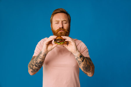 Bearded hungry man eating burger while standing isolated over blue backgroundの写真素材