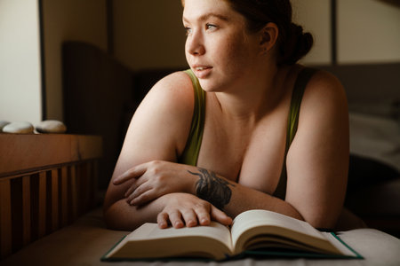 Ginger young woman wearing tank reading book while lying by window indoorsの写真素材