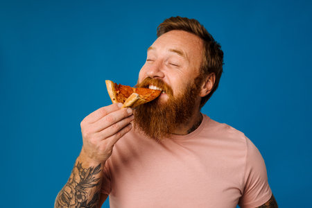 Bearded happy man eating pizza while standing isolated over blue backgroundの写真素材