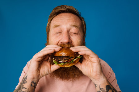 Bearded hungry man eating burger while standing isolated over blue backgroundの写真素材