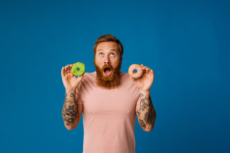 Bearded excited man holding donuts while standing isolated over blue backgroundの写真素材