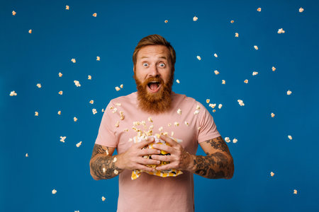 Shocked bearded white man in casual wear holding bucket with flying popcorn isolated over blue studio backgroundの写真素材