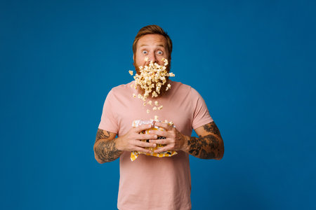 Shocked bearded white man in casual wear holding bucket with flying popcorn isolated over blue studio backgroundの写真素材