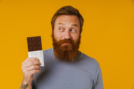 Bearded happy man holding chocolate bar while standing isolated over yellow backgroundの写真素材