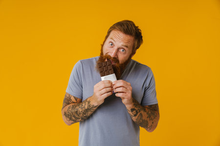 Bearded happy man eating chocolate bar while standing isolated over yellow backgroundの写真素材