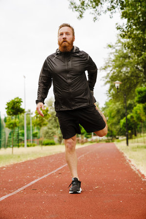 Adult handsome bearded redhead man stretching his leg looking aside, while standing on running track...
