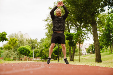 Adult handsome bearded redhead man jumping in the air with raised arms and looking aside, while...