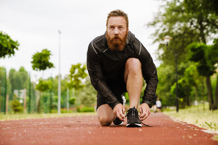Adult handsome bearded redhead man tying his shoelaces looking aside, while standing on his knee on running track outdoorsの写真素材