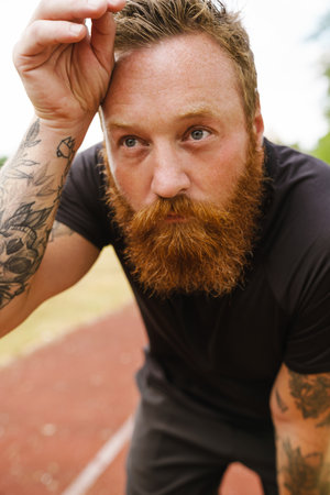 Portrait of adult bearded redhead man wiping sweat from forehead, leaning on his knee and looking aside, while standing on running track outdoorsの写真素材