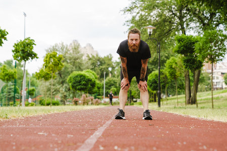 Portrait of adult bearded redhead man leaning on his knees and looking at camera, while standing on running track outdoorsの写真素材