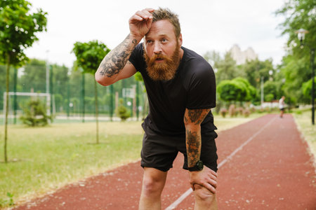 Portrait of adult bearded redhead man leaning on his knee, wiping sweat from forehead and looking aside, while standing on running track outdoorsの写真素材
