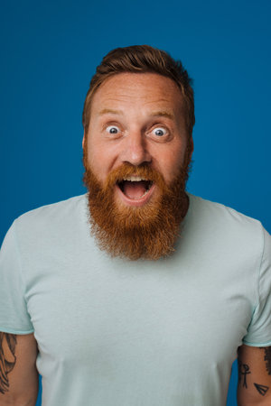 Adult bearded tattooed handsome stylish enthusiastic man with opened mouth, looking at camera , while standing over isolated blue backgroundの写真素材