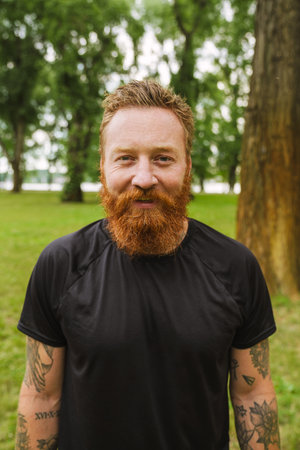 Portrait of young handsome bearded tattooed redhead smiling man looking at camera, while standing in green parkの写真素材