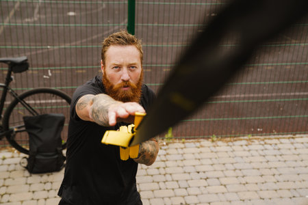 Young handsome tattooed bearded serious redhead man looking up and doing exercises with TRX near bicycle at outdoor sports fieldの写真素材