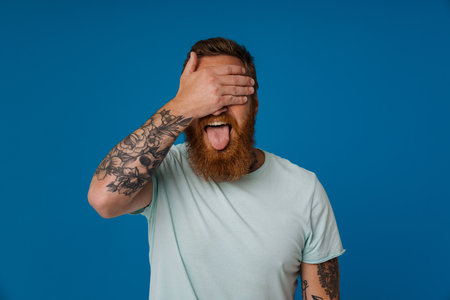 Portrait of adult bearded tattooed man showing tongue covering eyes with hand, while standing over isolated blue backgroundの写真素材