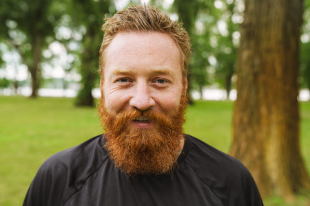Portrait of young handsome bearded redhead smiling man looking at camera, while standing in green parkの写真素材
