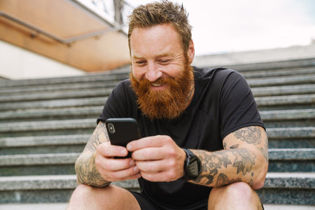 Young handsome bearded tattooed redhead smiling man holding and using his phone, while sitting on the stairs outdoorsの写真素材