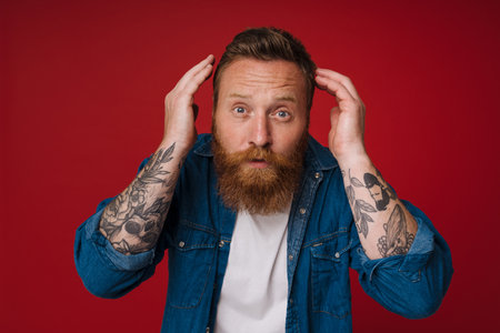 Adult bearded tattooed handsome stylish redhead man adjusting his haircut and looking at camera, while standing over isolated red backgroundの写真素材