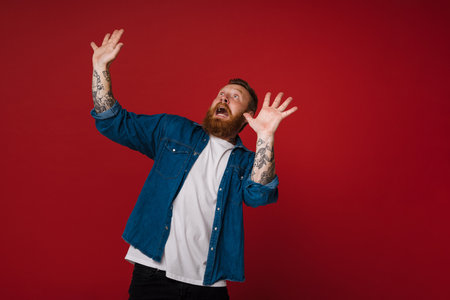 Adult bearded tattooed handsome scared redhead man with raised hands and opened mouth looking aside, while standing over isolated red backgroundの写真素材