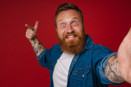 Selfie of adult bearded tattooed handsome enthusiastic redhead man doing gun gesture with raised hand and fingers looking at camera, while standing over isolated red backgroundの写真素材
