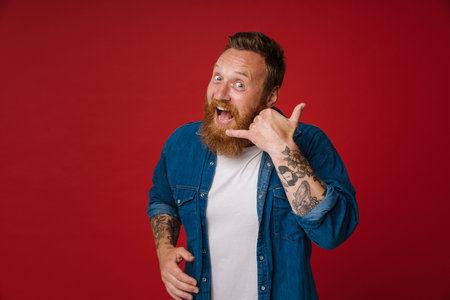 Adult redhead bearded tattooed enthusiastic man with opened mouth doing phone gesture and looking at camera, while standing over isolated red backgroundの写真素材