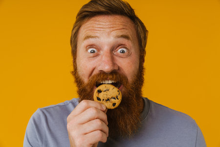 Bearded happy man smiling and eating cookies while standing isolated over yellow backgroundの写真素材