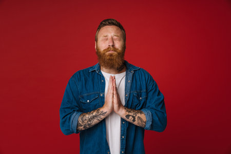Adult bearded tattooed handsome praying redhead man with folded hands and closed eyes standing over isolated red backgroundの写真素材