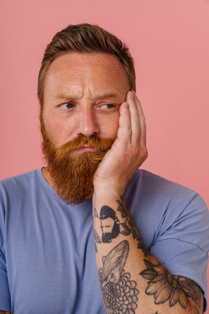 Ginger perplexed man with beard frowning and looking aside isolated over pink backgroundの写真素材