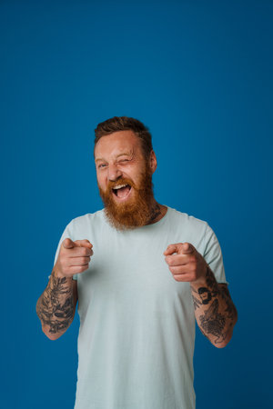 Ginger excited man winking and pointing fingers at camera isolated over blue backgroundの写真素材