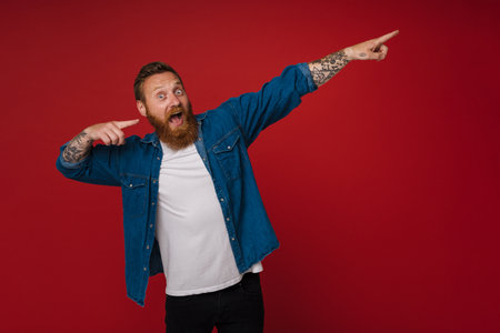 Ginger excited man with tattoo gesturing and pointing fingers aside isolated over red backgroundの写真素材
