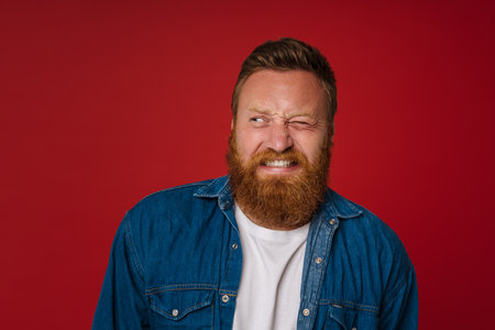 Ginger displeased man with tattoo grimacing and looking aside isolated over red backgroundの写真素材