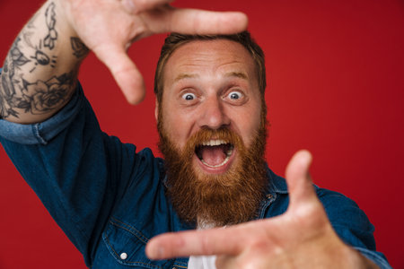 Ginger shocked man with tattoo making frame gesture isolated over red backgroundの写真素材