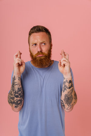 Ginger displeased man frowning and holding fingers crossed for best luck isolated over pink backgroundの写真素材