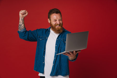 Ginger excited man wearing denim shirt gesturing and using laptop isolated over red backgroundの写真素材
