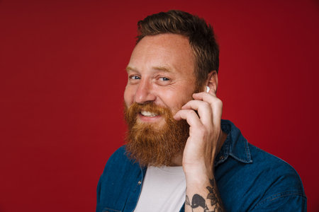 Ginger white man in earphones smiling and listening music isolated over red background