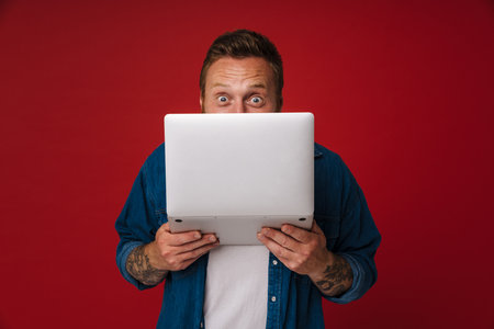 Ginger shocked man wearing denim shirt posing with laptop isolated over red backgroundの写真素材