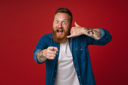 Ginger excited man smiling while making handset gesture isolated over red backgroundの写真素材