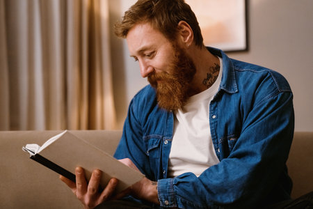 Middle-aged redhead bearded man reading book while sitting on sofa in cozy living room at homeの写真素材