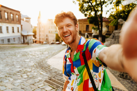 Selfie of young handsome smiling redhead man in colorful shirt standing on the streetの写真素材