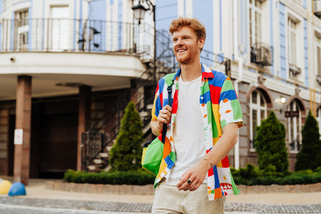 Young handsome redhead smiling man with stubble in colorful shirt walking street, near modern...