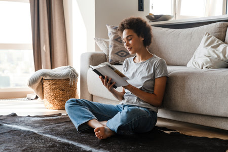 Young curly woman wearing jeans reading book while sitting on floor at homeの写真素材